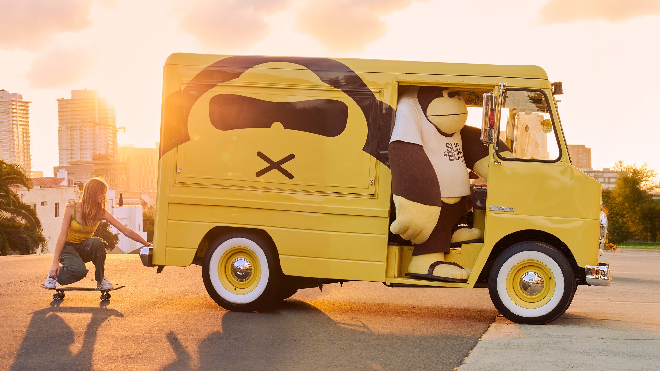 Girl skateboards behind yellow Sun Bum van with Sunny the mascot at sunset in the city – About Us Collection - Sun Bum.