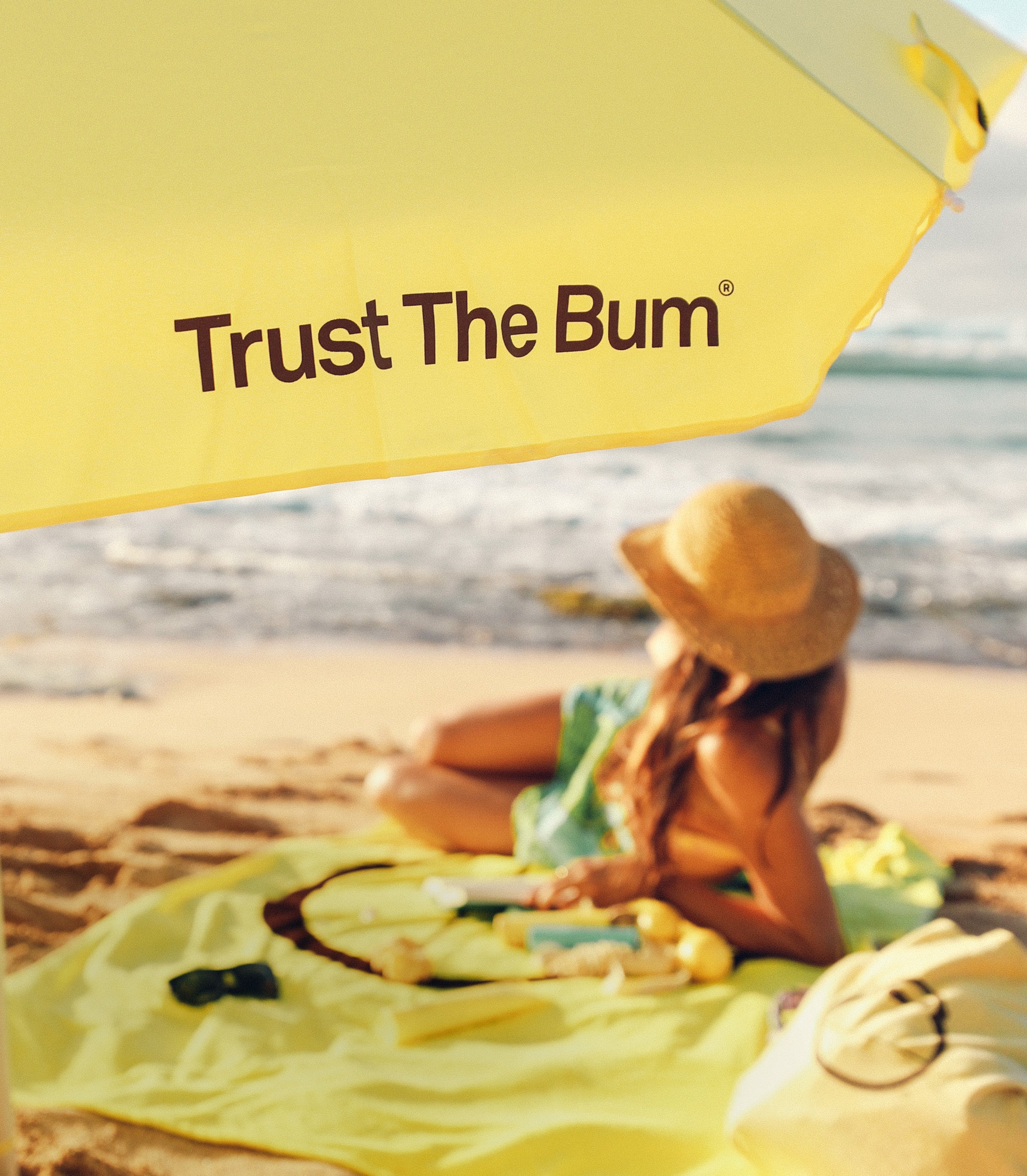 Woman lounging under a yellow “Trust The Bum” beach umbrella with Sun Bum essentials by the ocean – About Us Header - Sun Bum.