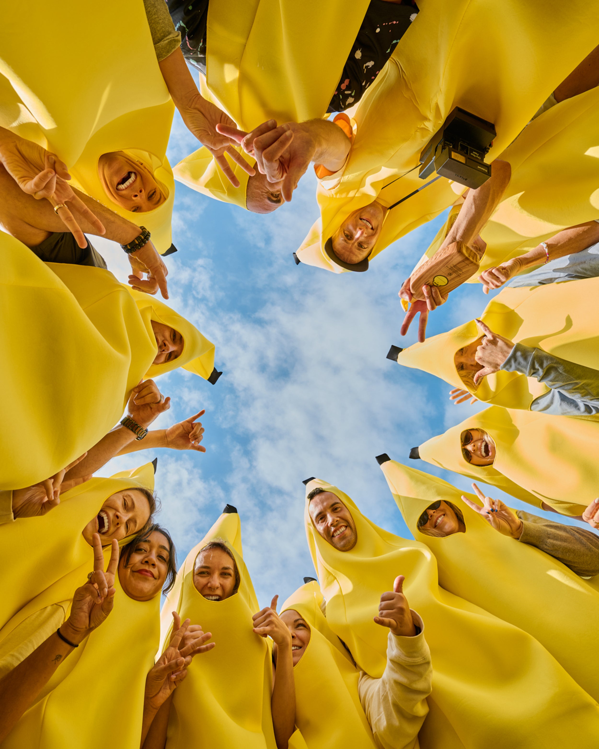 Team dressed in banana costumes smiles and throws peace signs in a circle under blue skies – Here's Our Story Collection - Sun Bum.