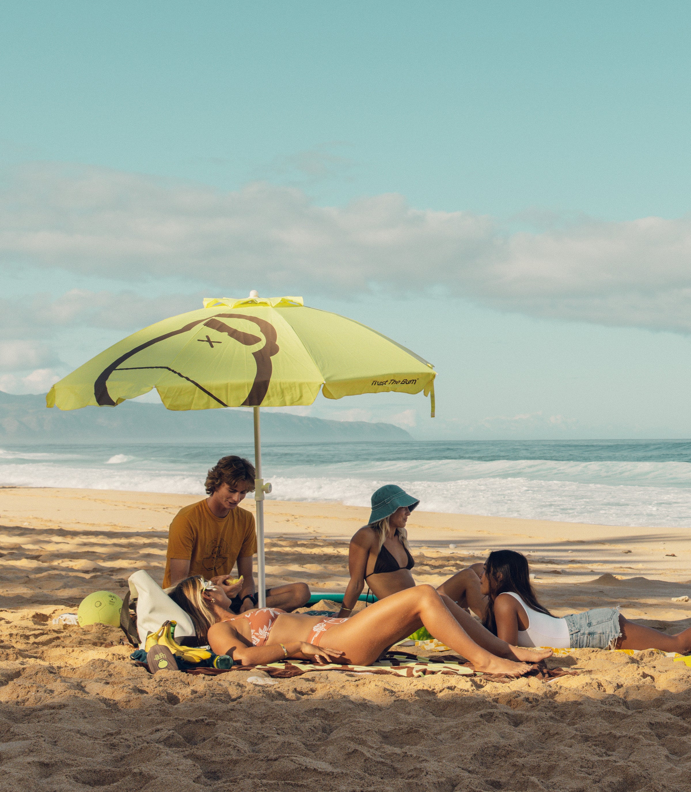 Group of friends relaxing under a Sun Bum umbrella on the sand, featuring the SB Beach Day Essentials Collection – Sun Bum.