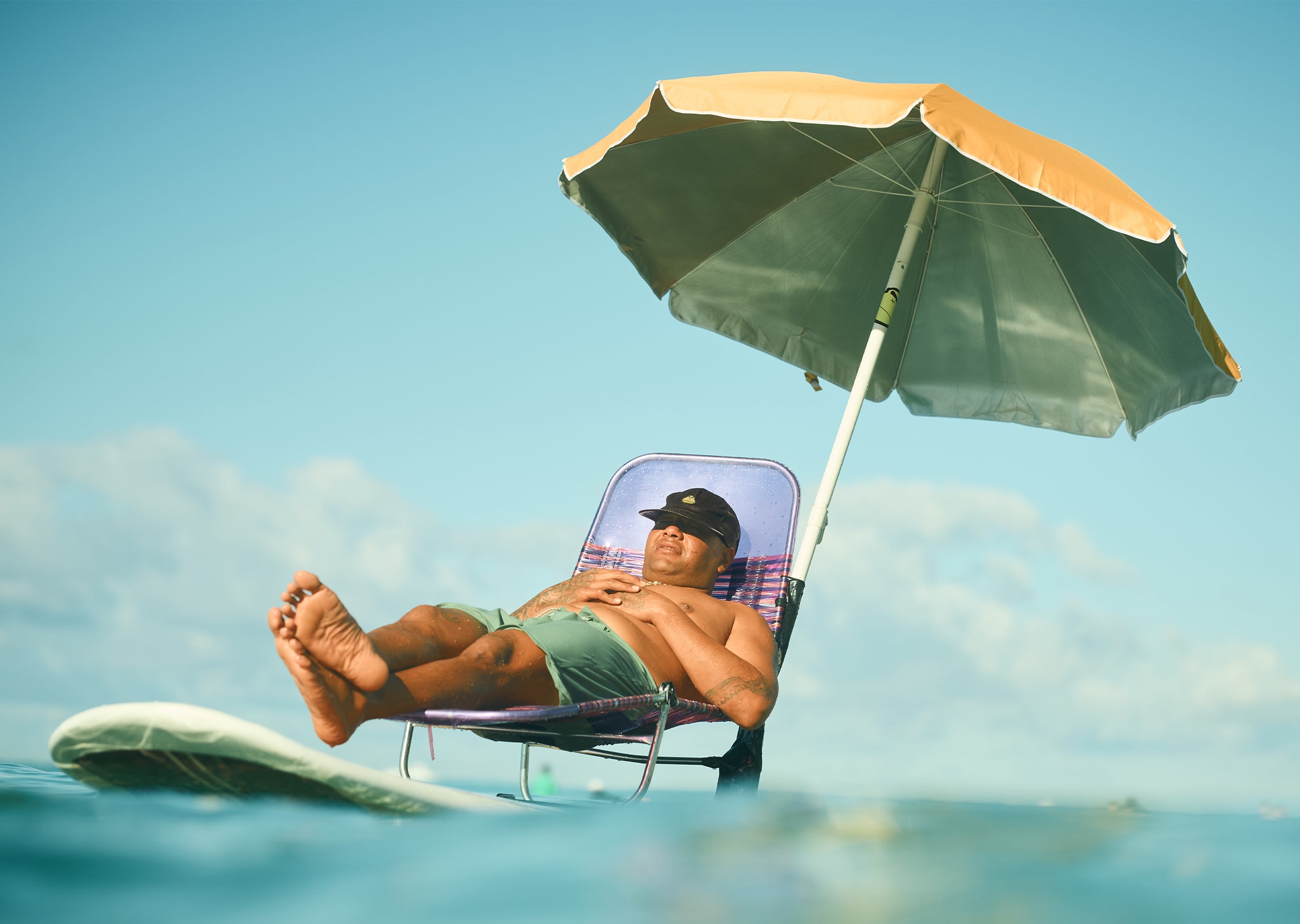 Man relaxing on a beach chair and umbrella balanced on a surfboard, showing creative sun protection from Know Your Spots Collection - Sun Bum.