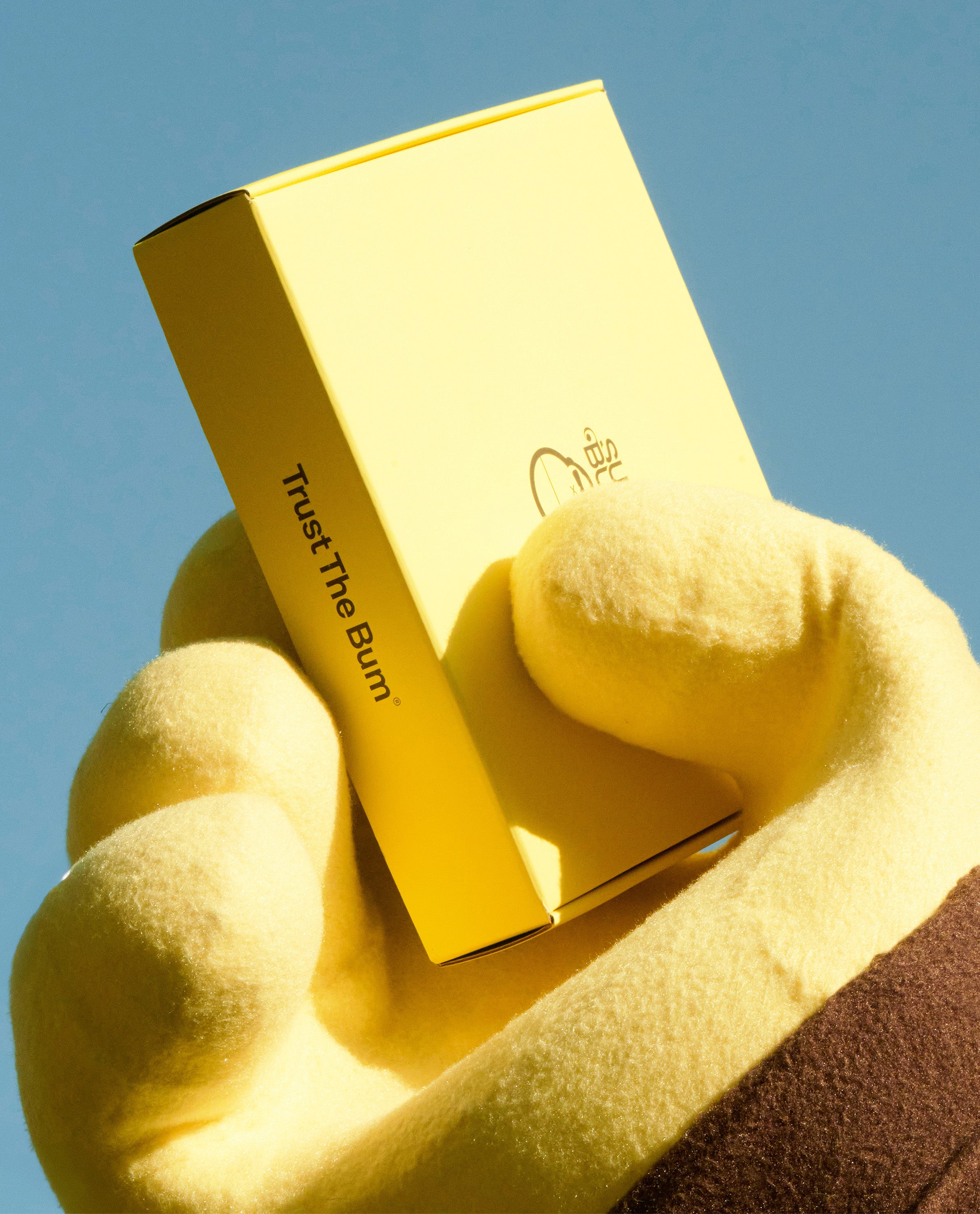 Hand holding a yellow product box with 'Trust The Bum' branding against a blue sky.