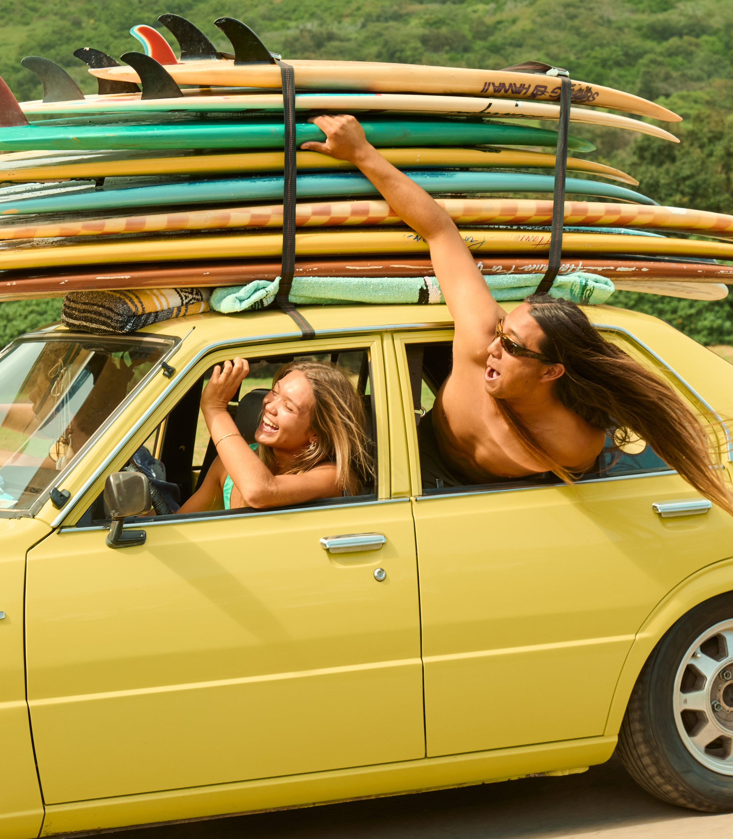 Car with a lot of surf boards strapped on top and people hanging out the windows.